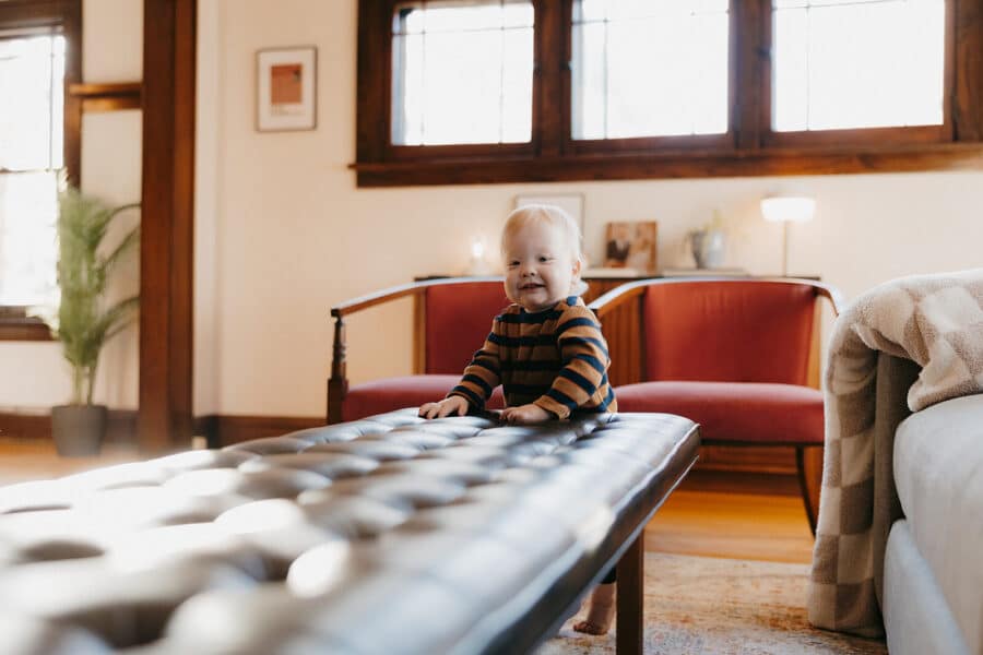 Baby smiling while playing on a leather ottoman in a cozy living room with modern decor and natural light.