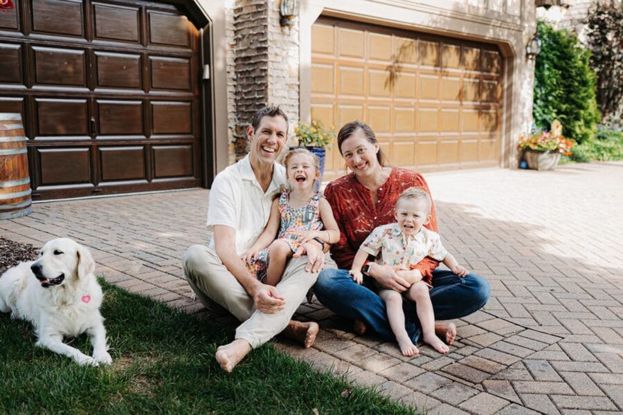 Family of four sitting on a driveway, smiling and laughing, with a golden retriever nearby, showcasing a joyful moment in a residential setting.