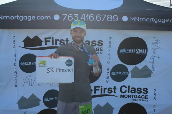 Runner holding a "5K Finisher" sign in front of a First Class Mortgage banner, celebrating participation in a community event.
