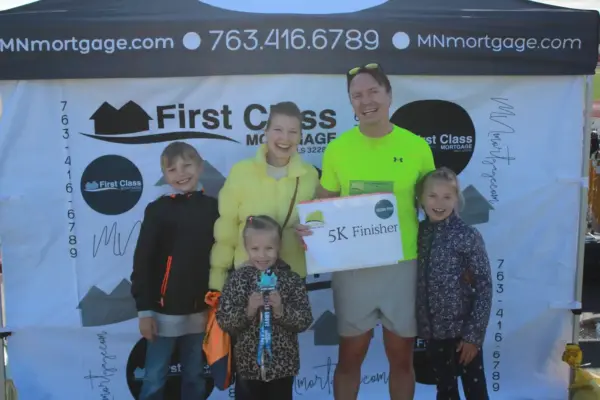 Group of five smiling individuals, including children, posing together holding a "5K Finisher" sign in front of a First Class Mortgage banner at a community event.
