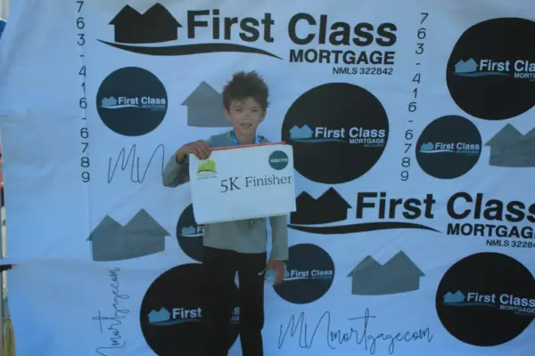 Child holding "5K Finisher" sign in front of First Class Mortgage backdrop, celebrating completion of a race event.