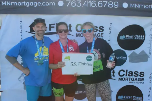 Three participants celebrating as 5K finishers, holding a sign, with First Class Mortgage branding in the background, at the Maple Grove Lions Half Marathon event.