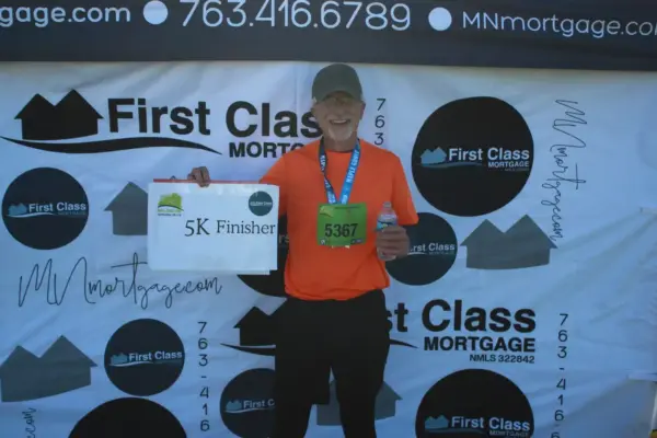 Participant holding a "5K Finisher" sign, wearing a medal and orange shirt, standing in front of a First Class Mortgage banner at a community race event.