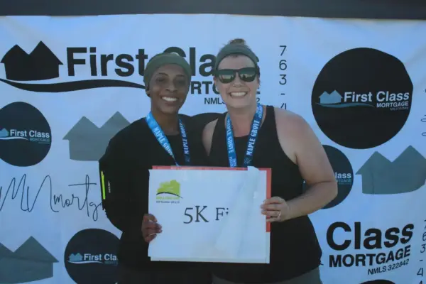 Two smiling women holding a "5K Finisher" sign, wearing medals, standing in front of a backdrop featuring the First Class Mortgage logo, celebrating their completion of a race.