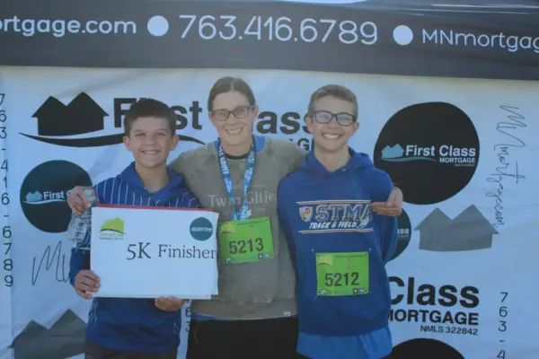 Three participants celebrating as 5K finishers, holding a sign, in front of a First Class Mortgage backdrop, showcasing community engagement in local events.