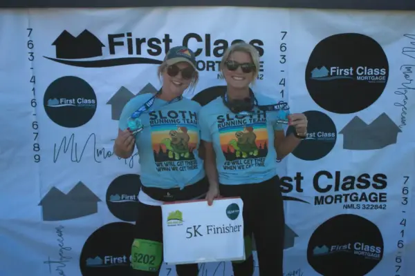 Two women wearing matching "Sloth Running Team" shirts, holding medals and a sign reading "5K Finisher," standing in front of a backdrop featuring the First Class Mortgage logo, celebrating their completion of a community race event.