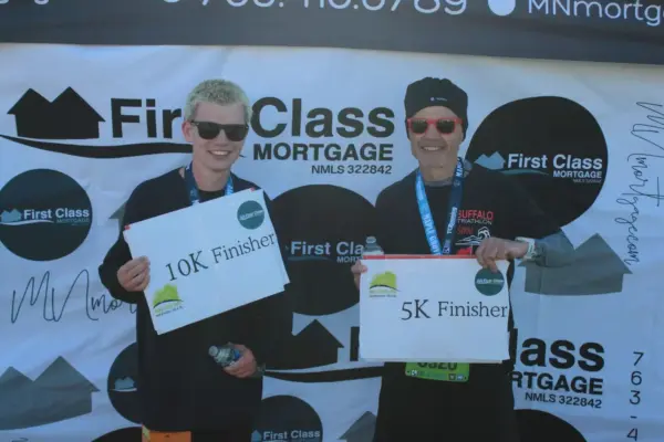 Two runners holding finisher signs, one for 10K and the other for 5K, celebrating their completion of a race sponsored by First Class Mortgage, with a branded backdrop featuring the company logo.