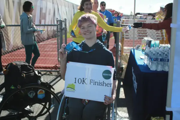 Participant in wheelchair holding "10K Finisher" sign and medal at Maple Grove Lions Half Marathon event, surrounded by cheering supporters and refreshments.