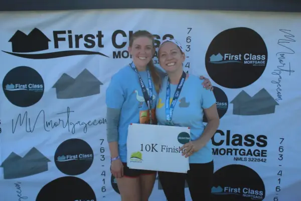 Two women smiling and holding a sign that reads "10K Finish," wearing medals and blue shirts, in front of a backdrop featuring the First Class Mortgage logo, celebrating a running event.