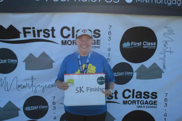 Participant holding "5K Finished" sign with medal, smiling in front of First Class Mortgage promotional backdrop, celebrating completion of Maple Grove Lions 5K race.