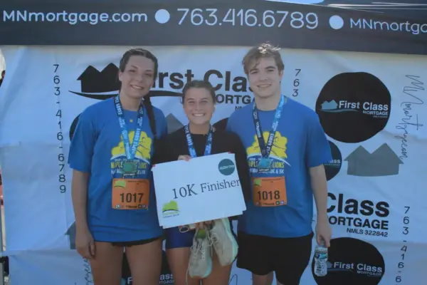 Participants celebrating their completion of the 10K race, wearing medals and blue shirts, holding a sign that reads "10K Finisher," in front of a First Class Mortgage banner at a community event.
