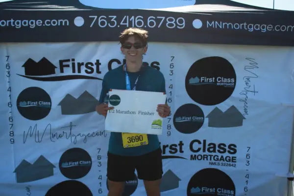 Young male runner holding a "Half Marathon Finisher" sign, standing in front of a branded backdrop featuring First Class Mortgage logos, celebrating completion of a race event.