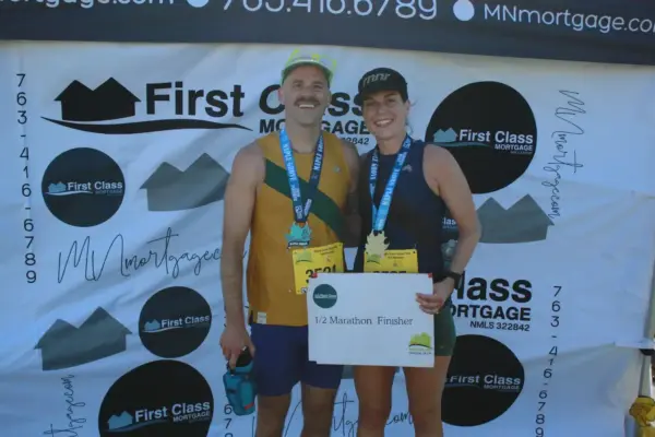 Runners celebrating as half marathon finishers, holding a sign, with First Class Mortgage branding in the background.