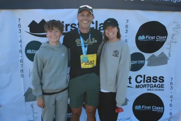 Participants celebrating at the Maple Grove Lions Half Marathon with a medal, posing in front of a First Class Mortgage banner.