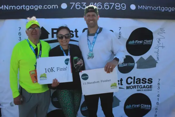 Participants celebrating their race achievements at the First Class Mortgage booth, holding signs for "10K Finisher" and "1/2 Marathon Finisher," showcasing community engagement in the Maple Grove Lions Half Marathon event.