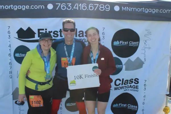 Three runners celebrating their 10K finish, holding a sign that reads "10K Finished," with a backdrop featuring the First Class Mortgage logo and contact information.