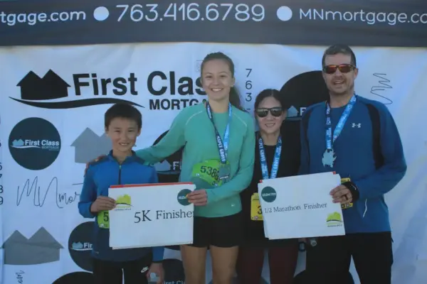 Participants celebrating their achievements at the First Class Mortgage-sponsored race, holding "5K Finisher" and "1/2 Marathon Finisher" signs, with the company logo in the background.