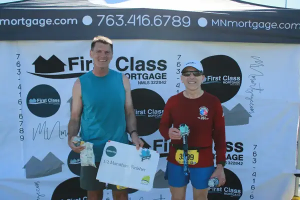 Two runners smiling post-race, holding a finisher's certificate, in front of a First Class Mortgage banner at a community event.
