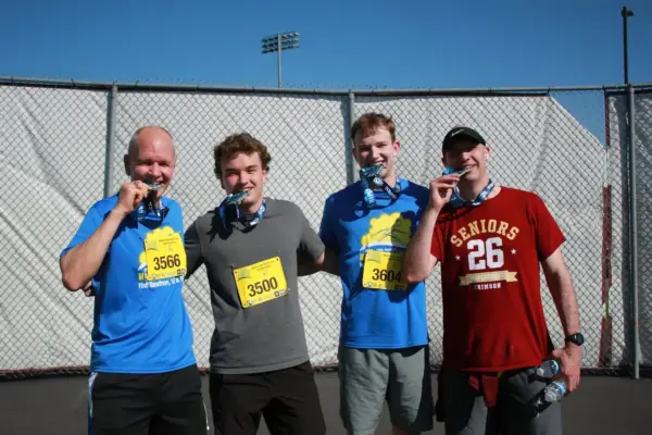 Four male runners celebrating with medals after finishing the Maple Grove Lions Half Marathon, wearing race bibs numbered 3566 and 3500, against a sports field backdrop.