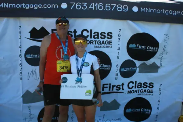 Participants celebrating as half marathon finishers, wearing medals, posing in front of a First Class Mortgage banner at the Maple Grove Lions Half Marathon event.