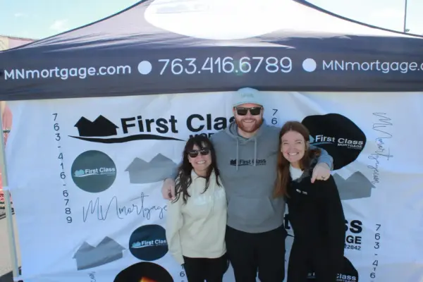 Three smiling individuals posing in front of a branded First Class Mortgage tent at a community event, showcasing engagement and support for local initiatives.