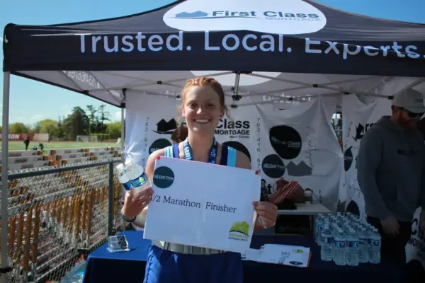 Smiling female runner holding a "Half Marathon Finisher" sign, standing in front of a First Class Mortgage tent at a community race event, with water bottles and promotional materials visible.