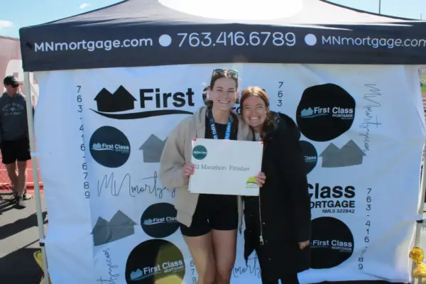 Participants celebrating at the Maple Grove Lions Half Marathon, holding a finisher's sign in front of the First Class Mortgage booth, showcasing community engagement and event sponsorship.