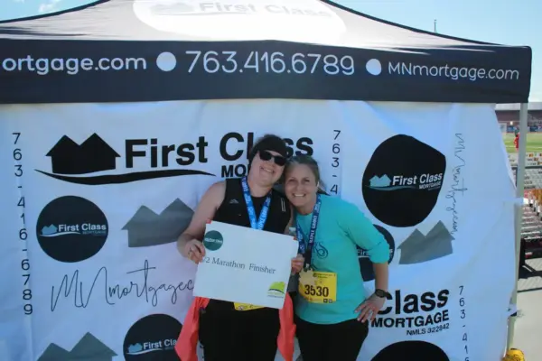 Two women celebrating at the First Class Mortgage booth, one holding a sign that says "12 Marathon Finisher," both wearing race medals, with a branded backdrop promoting First Class Mortgage.