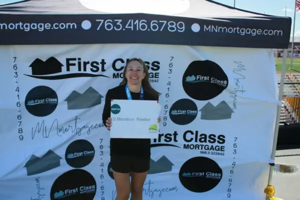 Woman holding a finisher sign at the First Class Mortgage booth after completing a marathon, with branding and contact information visible in the background.
