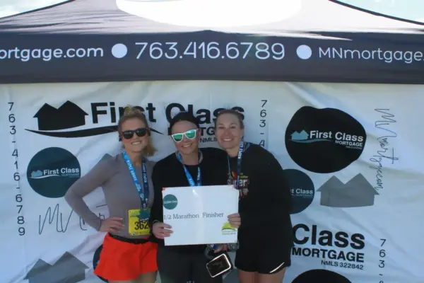 Three women celebrating as half marathon finishers in front of a First Class Mortgage banner, showcasing medals and a sign.