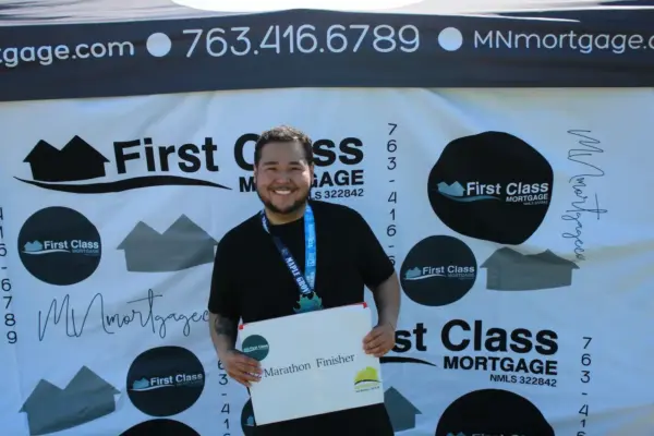 Smiling marathon finisher holding a sign in front of a First Class Mortgage backdrop, showcasing community engagement at the Maple Grove Lions Half Marathon.