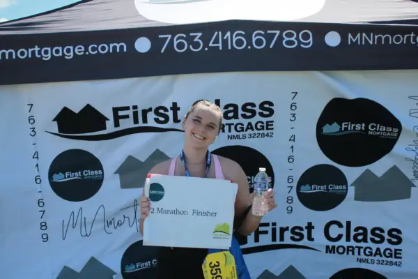 Woman holding a sign that reads "Half Marathon Finisher" and a water bottle, standing in front of a First Class Mortgage banner at a community event.