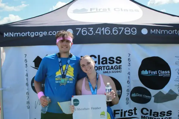 Two smiling participants holding a sign that reads "Half Marathon Finisher" in front of a First Class Mortgage tent, showcasing community engagement at the Maple Grove Lions Half Marathon event.